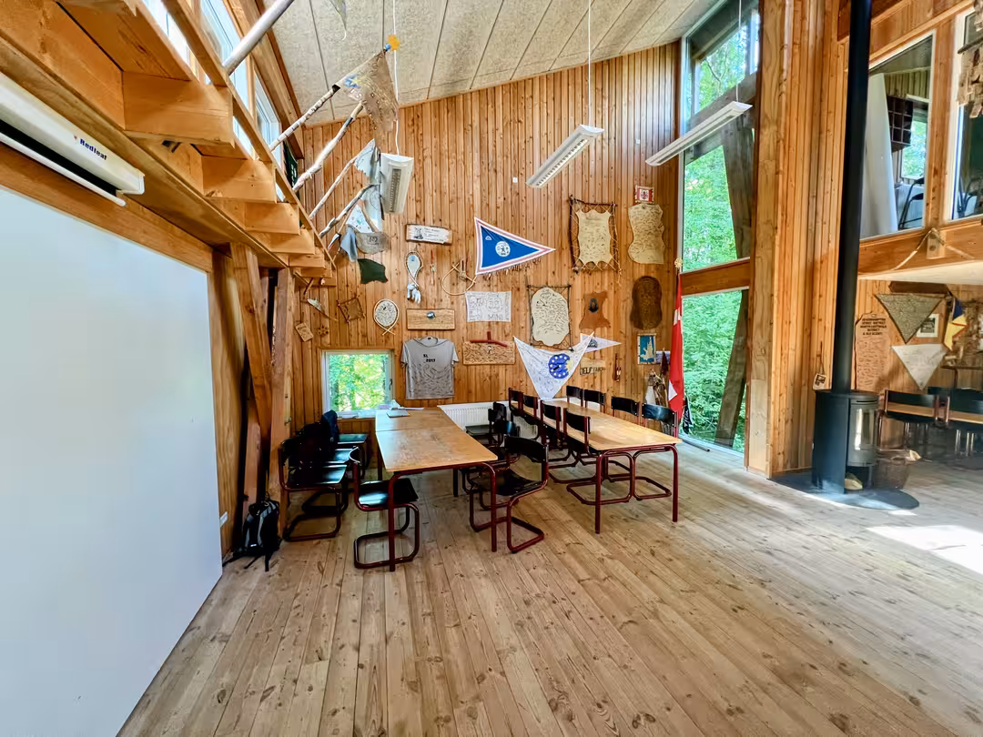Fællesrum i Bjørnholthytten med borde, stole og udsigt til skoven