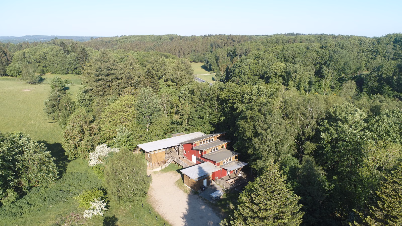 Luftfoto af Bjørnholthytten omgivet af skov og marker - set fra droneperspektiv